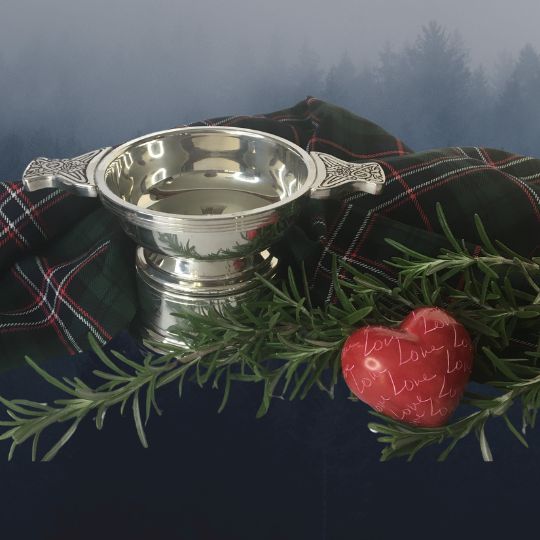 A silver quaich with
                        a sprig of rosemary, and red heart-shaped stone
                        that has the word Love repeated over its surface
                        and a length of the Scottish National Tartan, on
                        a white background