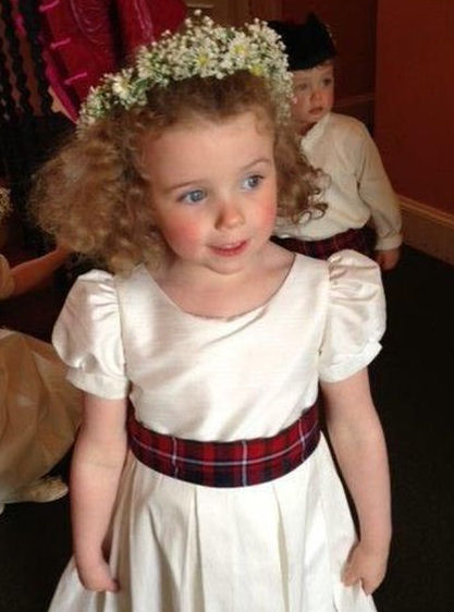 A flower girl wearing a white dress with a
                      tartan sash and a wreath of baby's breath on her
                      head