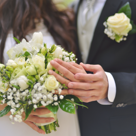 Close up of
                      bride and groom's hands. Groom holding the bride's
                      hand, bride holding a bouquet of white roses and
                      lily of the valley. The groom wears a dark suit, a
                      silver gray tie and has a white rose boutonniere
                      on his left lapel