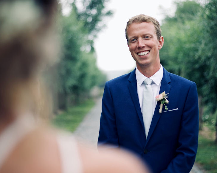 Bridegroom wearing
                        blue suit smiling at out-of-focus bride