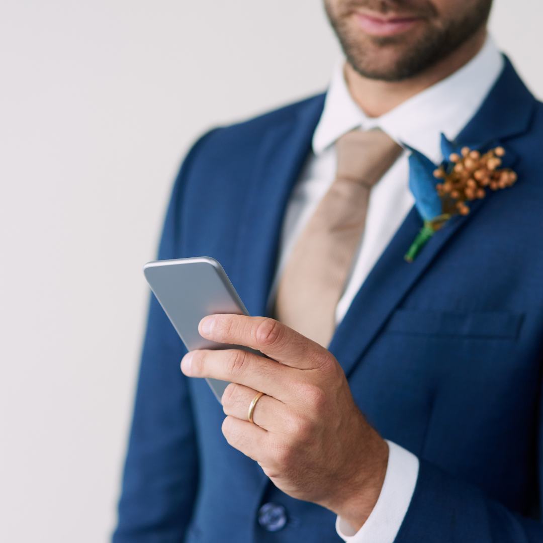 Groom
                      wearing a blue jacket on which is pinned a floral
                      boutonnier, is looking at the screen of a silver
                      smartphone