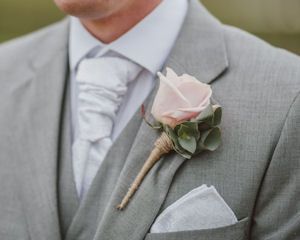 Man wearing a grey
                        suit with a pink rose pinned to the lapel and a
                        white pocket square in the breast pocket